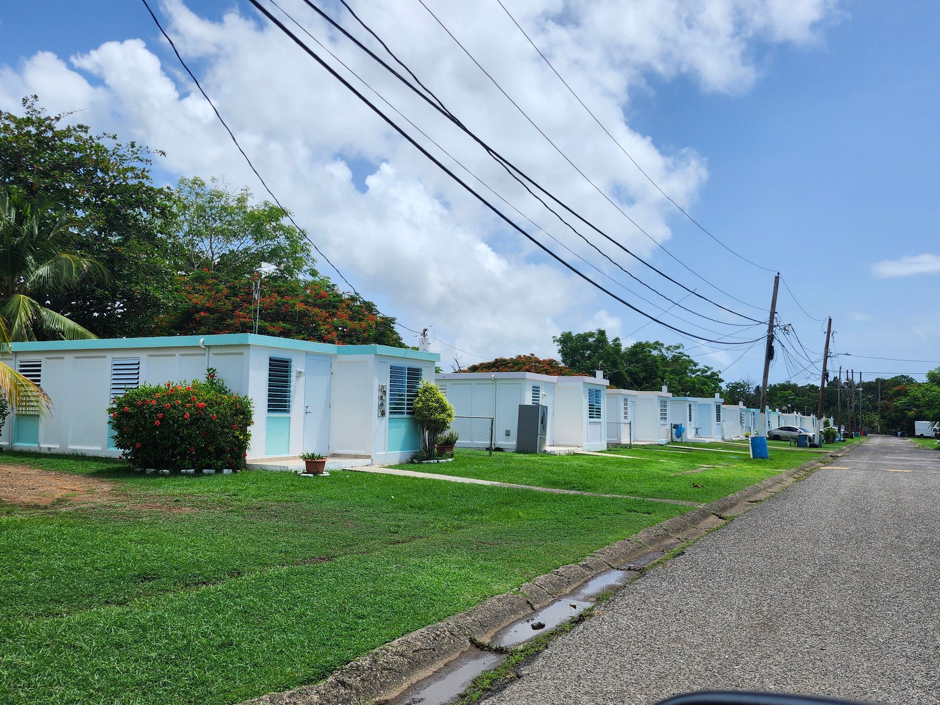 Small white houses along a gravel road under power lines, beside a green lawn and cloudy sky.