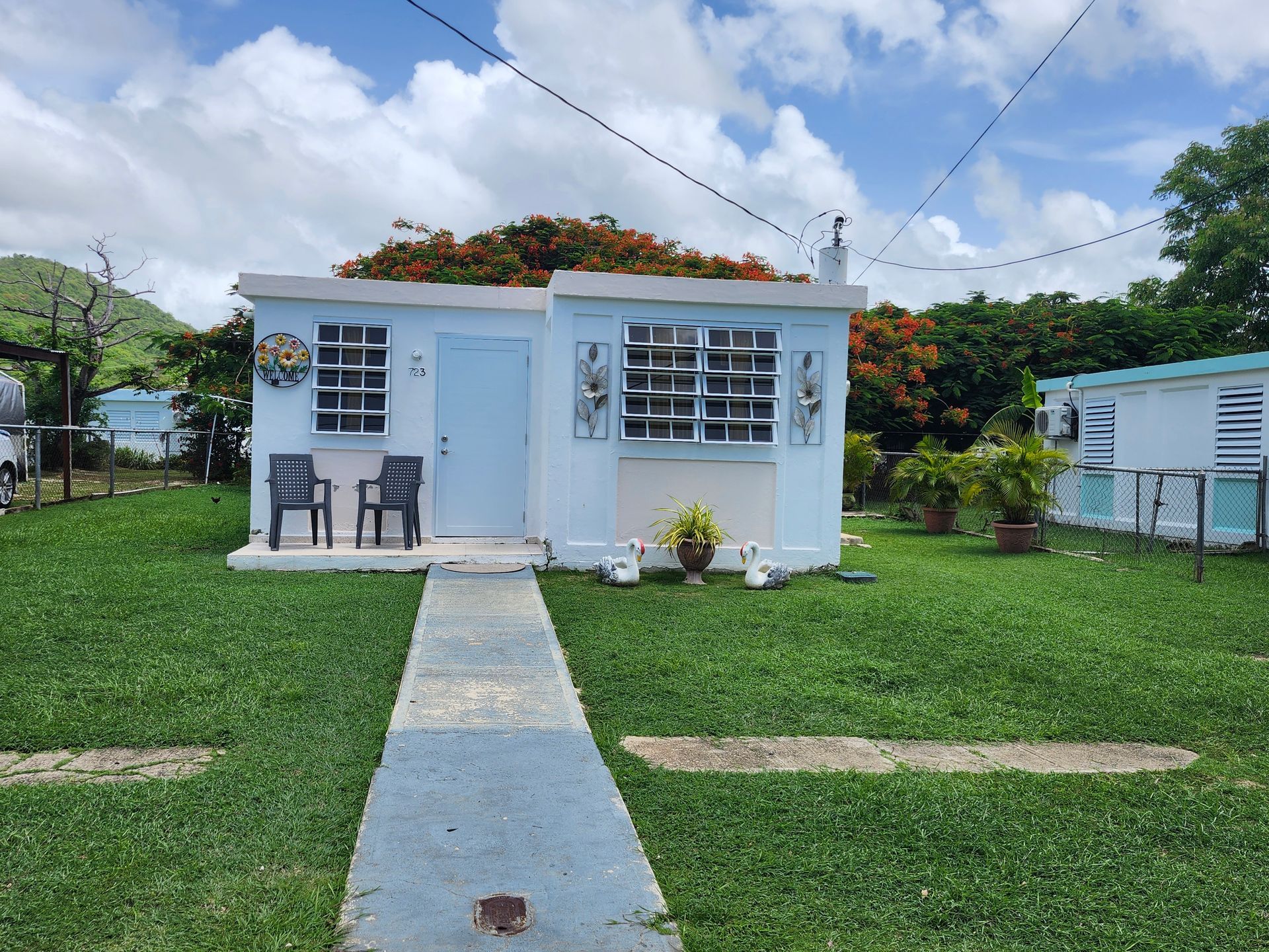 Small white cottage with blue trim, front walkway, and two chairs on a green lawn under a partly cloudy sky