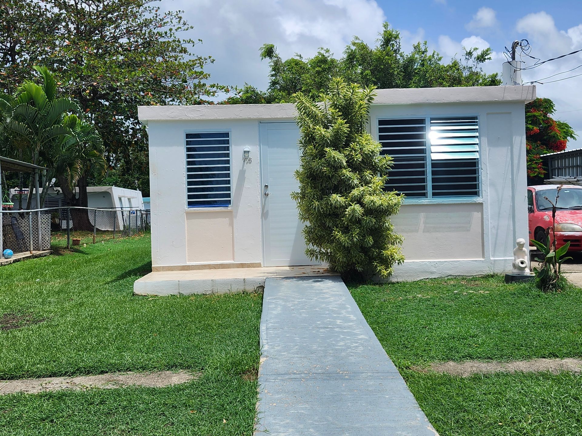 Small white building with blue shutters, a concrete path, and green grass under a sunny sky