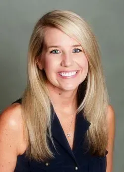 Woman with blonde hair smiling, wearing a navy top, against a gray background.