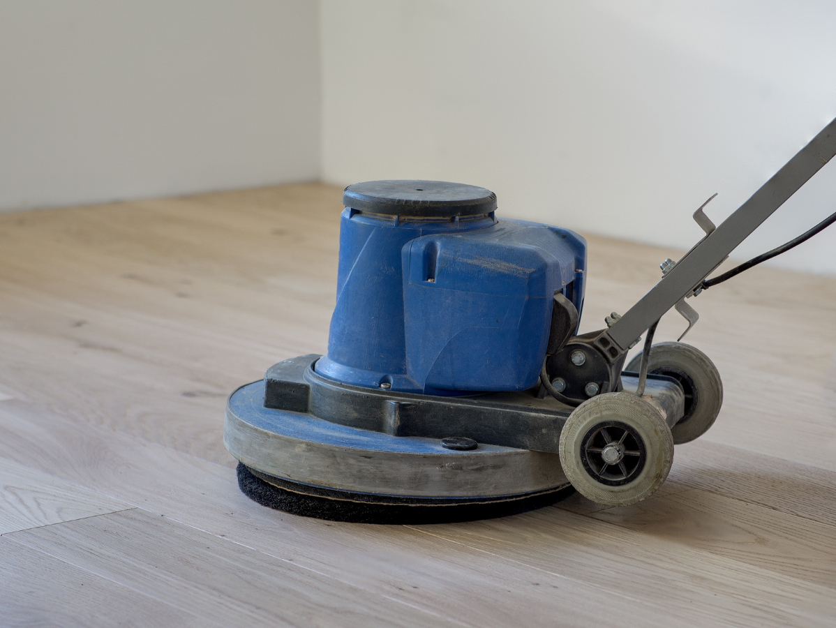 Blue floor buffer on a hardwood floor, with a person’s hands holding the handle.