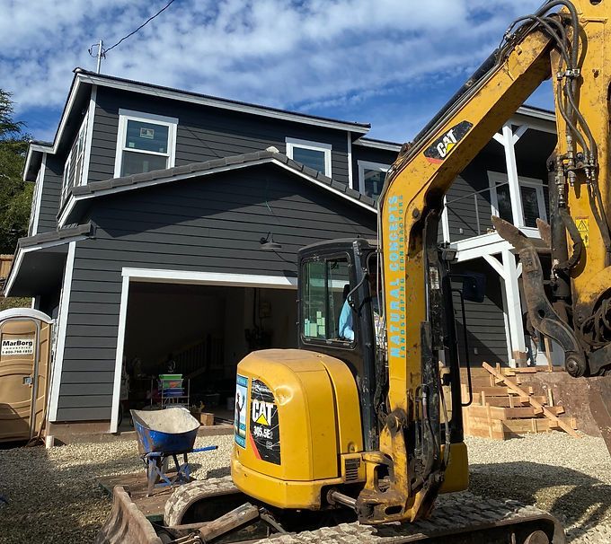 Yellow excavator in front of a black two-story house under construction, with an open garage.
