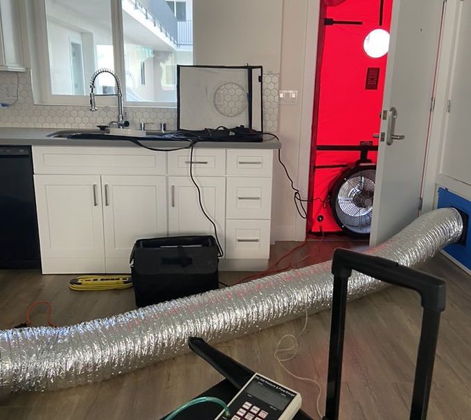 Kitchen with white cabinets, black box on floor, and silver ducting hose across the room near a red doorway.