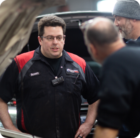 Man in black and red work jacket speaking beside an open car hood with two others nearby | Browncroft Garage