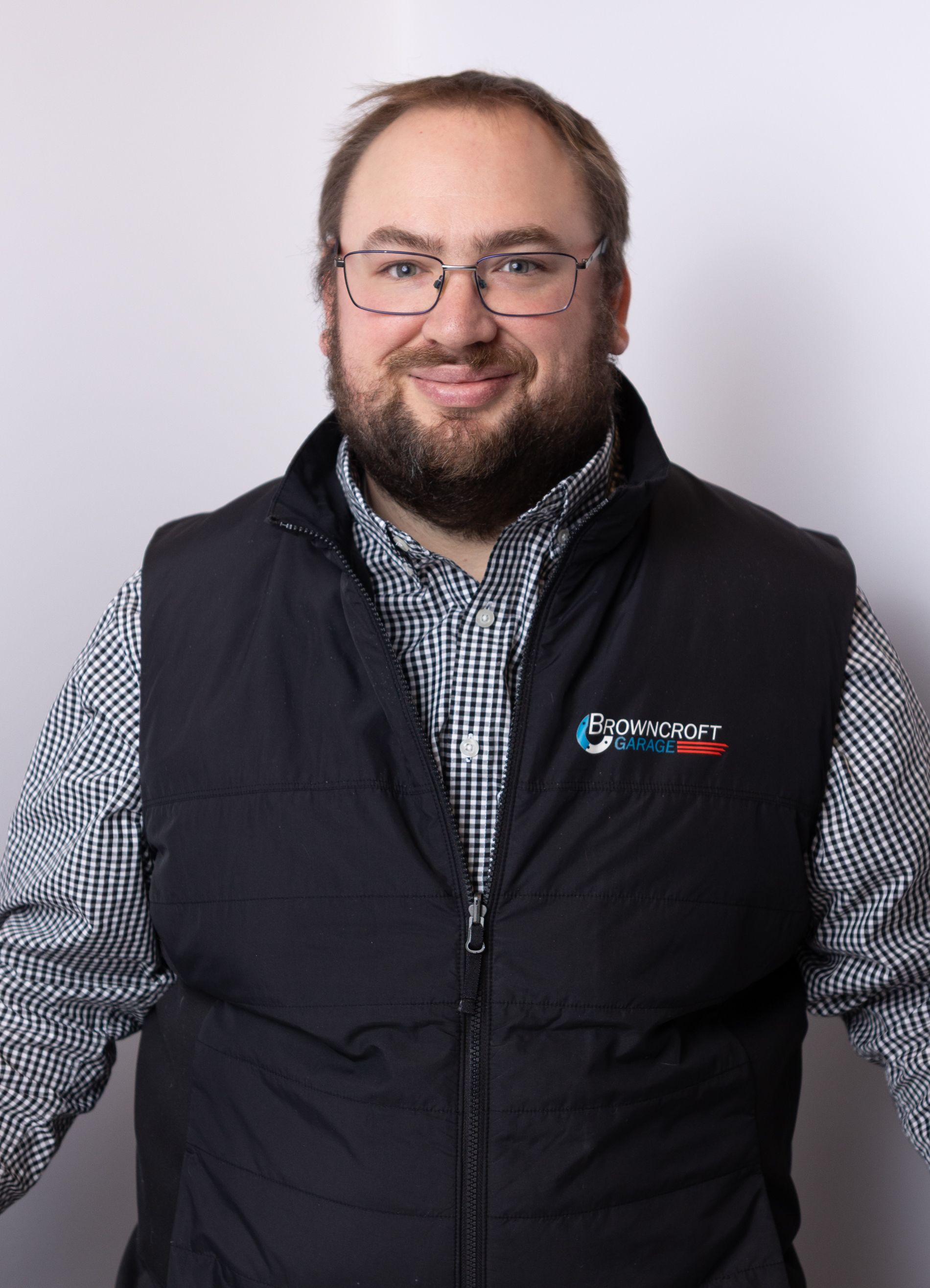 Smiling person wearing glasses and a black vest over a checkered shirt against a light background | Browncroft Garage