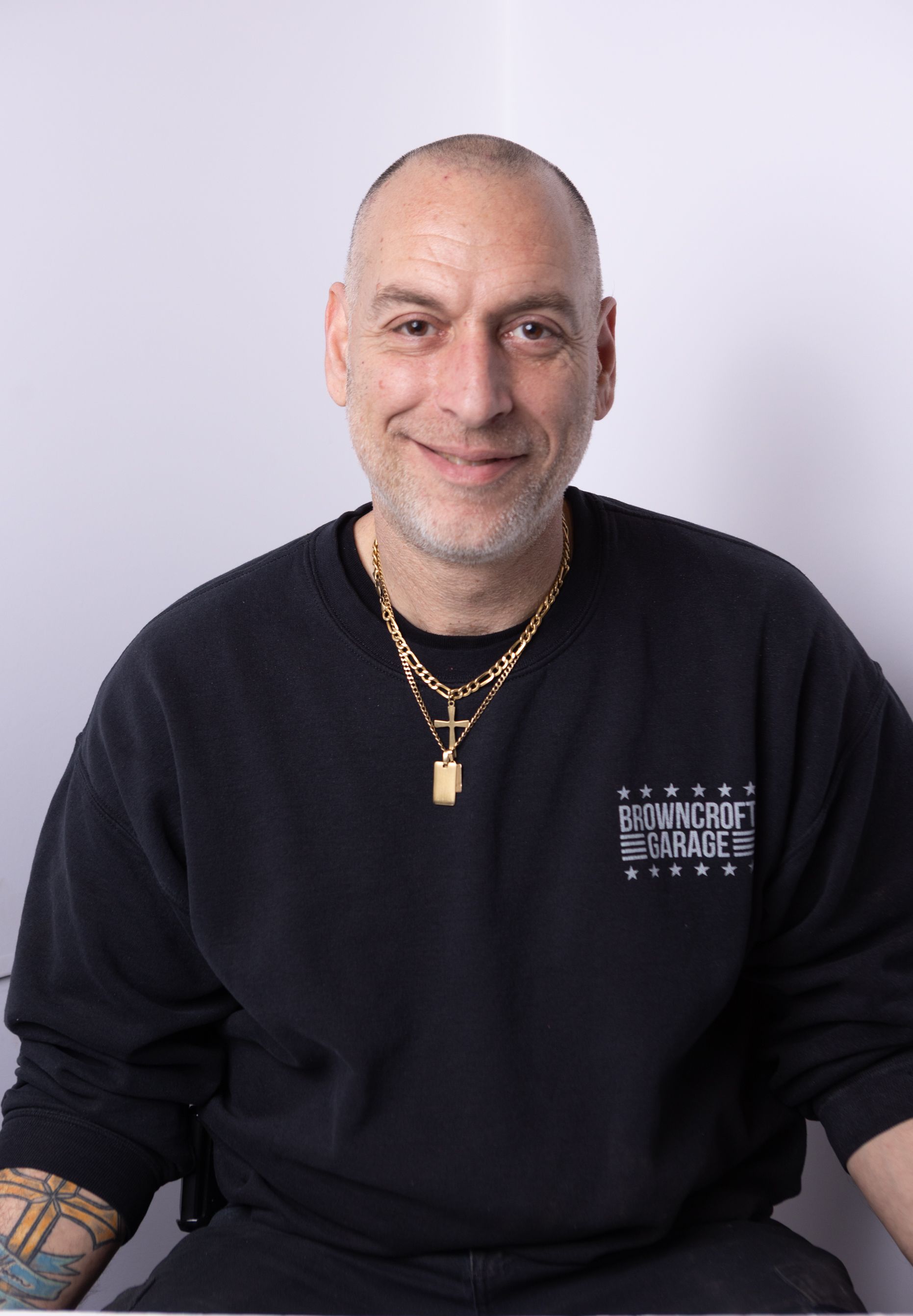 Man in a black sweatshirt with gold necklaces, smiling against a light background. | Browncroft Garage