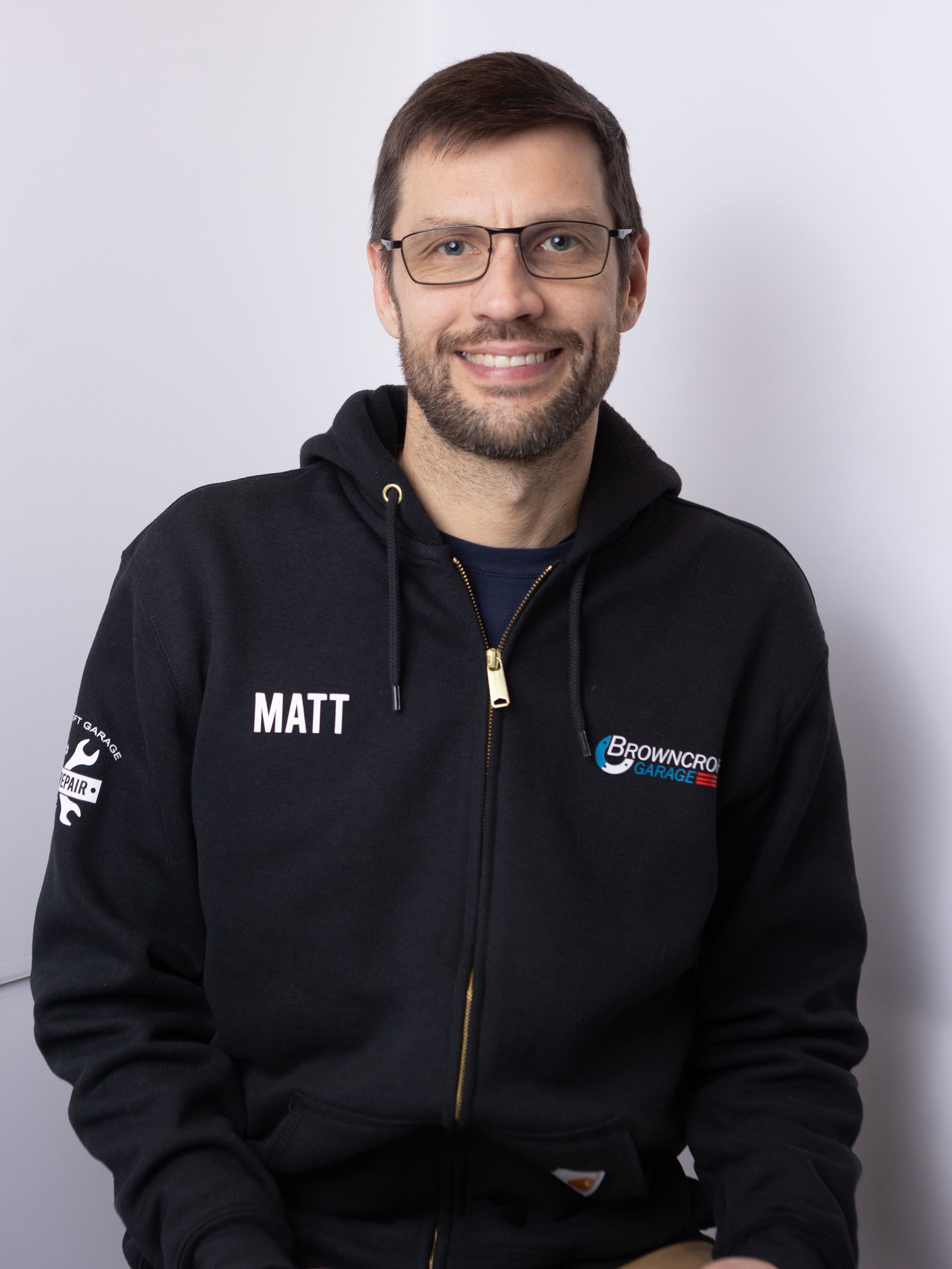 Smiling person in a black hoodie with glasses, seated against a light gray background. | Browncroft Garage