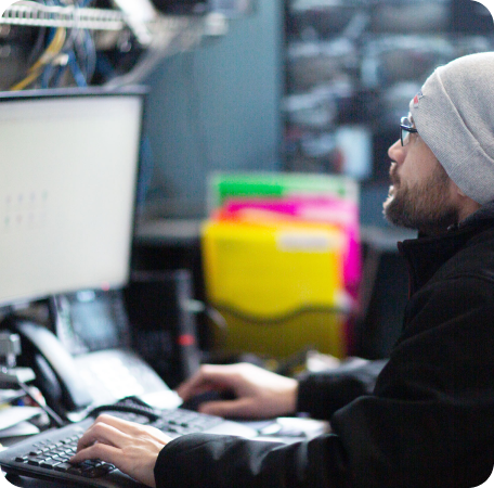 Person typing at a desktop computer in an office, wearing a gray beanie and black jacket | Browncroft Garage