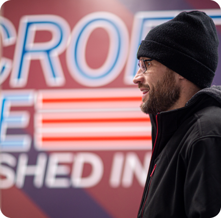 Profile of a bearded man in a black beanie and glasses beside a neon sign | Browncroft Garage