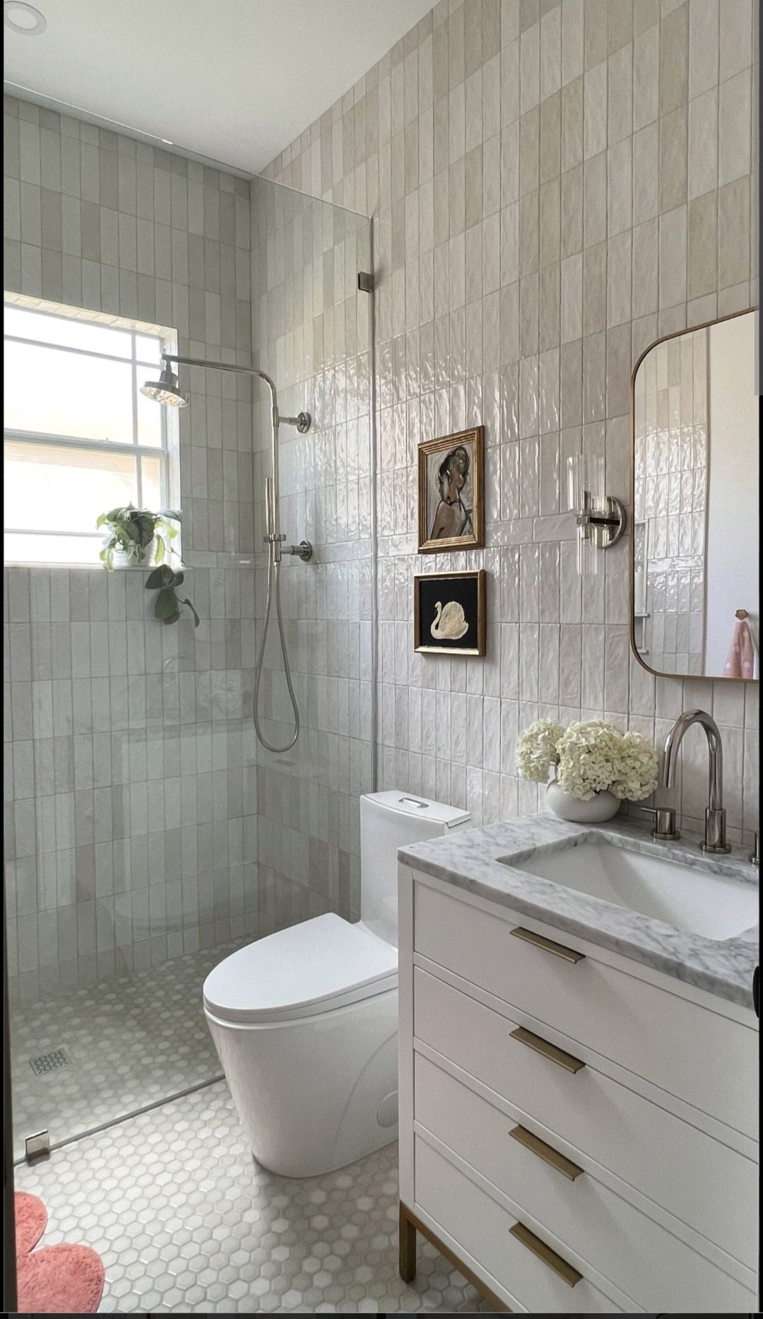 A modern bathroom featuring a white vanity with gold hardware, a toilet, a glass shower, and light-toned vertical tiles.