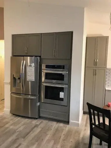 A stainless steel refrigerator and built-in oven set next to a tall pantry cabinet in a kitchen with light wood floors.
