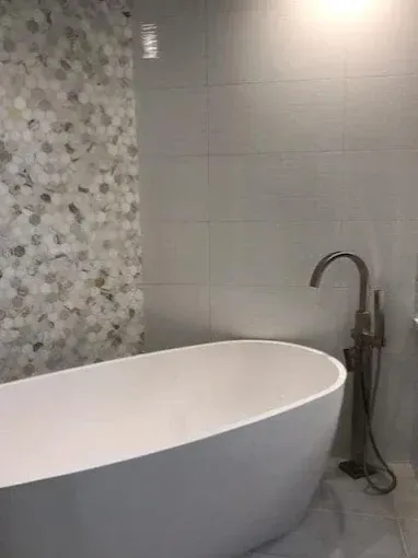 A white freestanding bathtub stands next to a bronze floor-mounted faucet in front of a gray tiled wall.