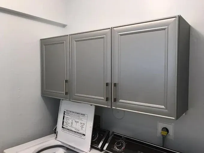 Three gray kitchen-style cabinets mounted on a white wall above a laundry machine.