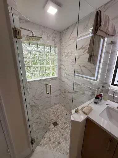 A modern bathroom with a glass-enclosed shower, marble-patterned tile walls, a pebble-stone floor, and a wooden vanity.