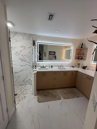 A modern bathroom features a double vanity with light wood cabinets, white counters, an LED mirror, and a tiled shower.