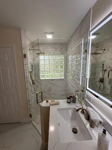 A modern bathroom featuring a white marble vanity, a mirror with integrated LED lighting, and a walk-in glass shower.