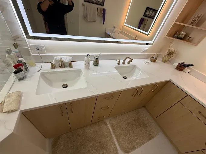 A modern double-sink bathroom vanity with light wood cabinets, white marble countertops, and an illuminated mirror.