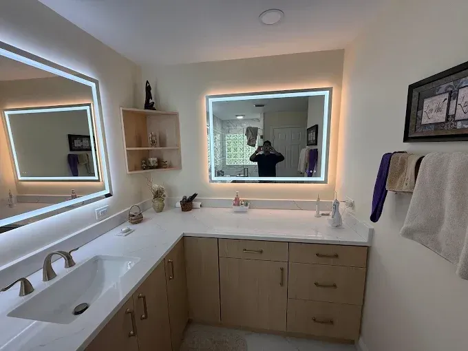 A brightly lit bathroom with two mirrors, light wood cabinetry, white countertops, and a white sink.