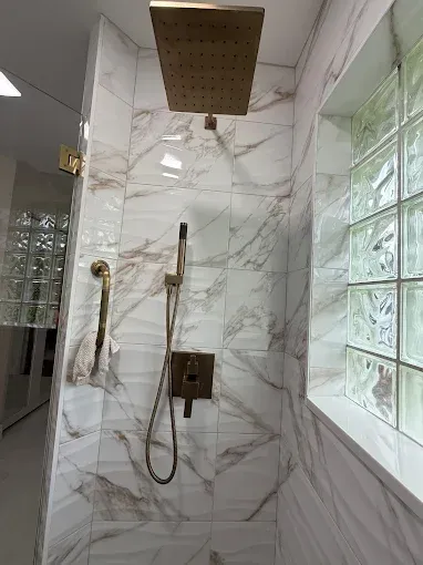 A walk-in shower with gold-toned fixtures, a square rainfall showerhead, a handheld sprayer, and white marble-look tiles.