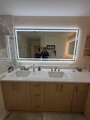 A double-sink bathroom vanity with light wood cabinets, white countertops, and a large, rectangular illuminated mirror.