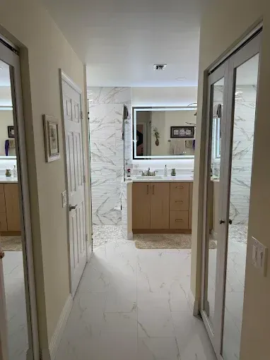 A hallway view of a bathroom with white marble-patterned tile, a light wood vanity, and mirrored closet doors on each side.