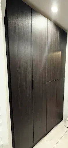 A tall, dark brown wooden wardrobe with vertical grain and sleek, integrated handles against a plain white wall.