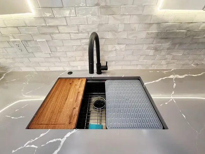 A modern kitchen sink with a matte black faucet, a wooden cutting board, and a gray drying rack set into marble counters.
