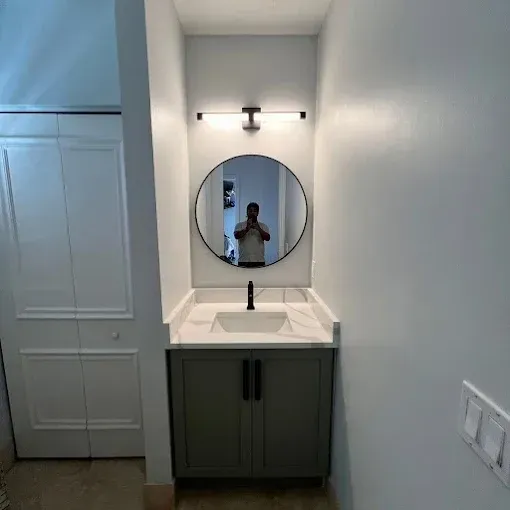 A modern bathroom vanity with a grey cabinet, white countertop, circular mirror, and wall-mounted light fixture.