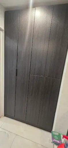 A tall, dark brown wardrobe with vertical paneling and sleek handles, situated against a white wall on a tiled floor.