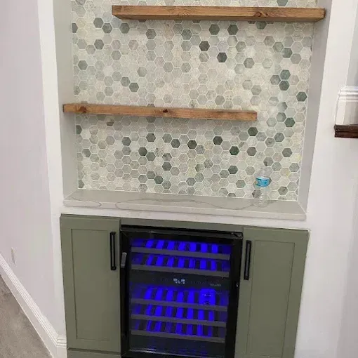 A built-in wet bar with a wine cooler, sage green cabinets, hexagon tile backsplash, and two wooden floating shelves.