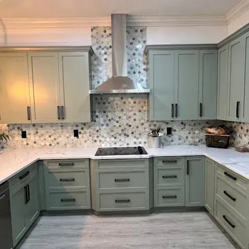 A modern kitchen with sage green cabinets, white marble countertops, a hexagonal patterned backsplash, and steel hood.
