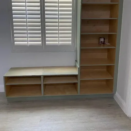 Custom-built light-wood shelving unit and bench seat installed beneath a window with white plantation shutters.