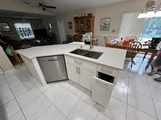 A white kitchen island with a stainless steel dishwasher, double sink, and an open cabinet drawer in a tiled kitchen.