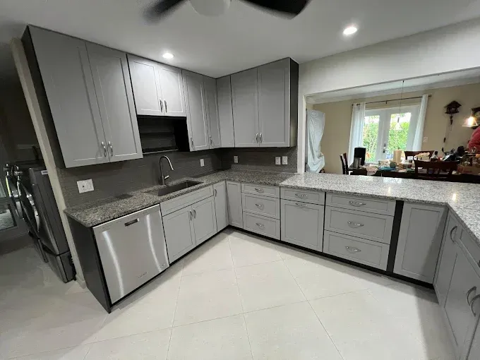 A kitchen with light gray cabinets, granite countertops, a stainless steel dishwasher, and an opening to a dining area.
