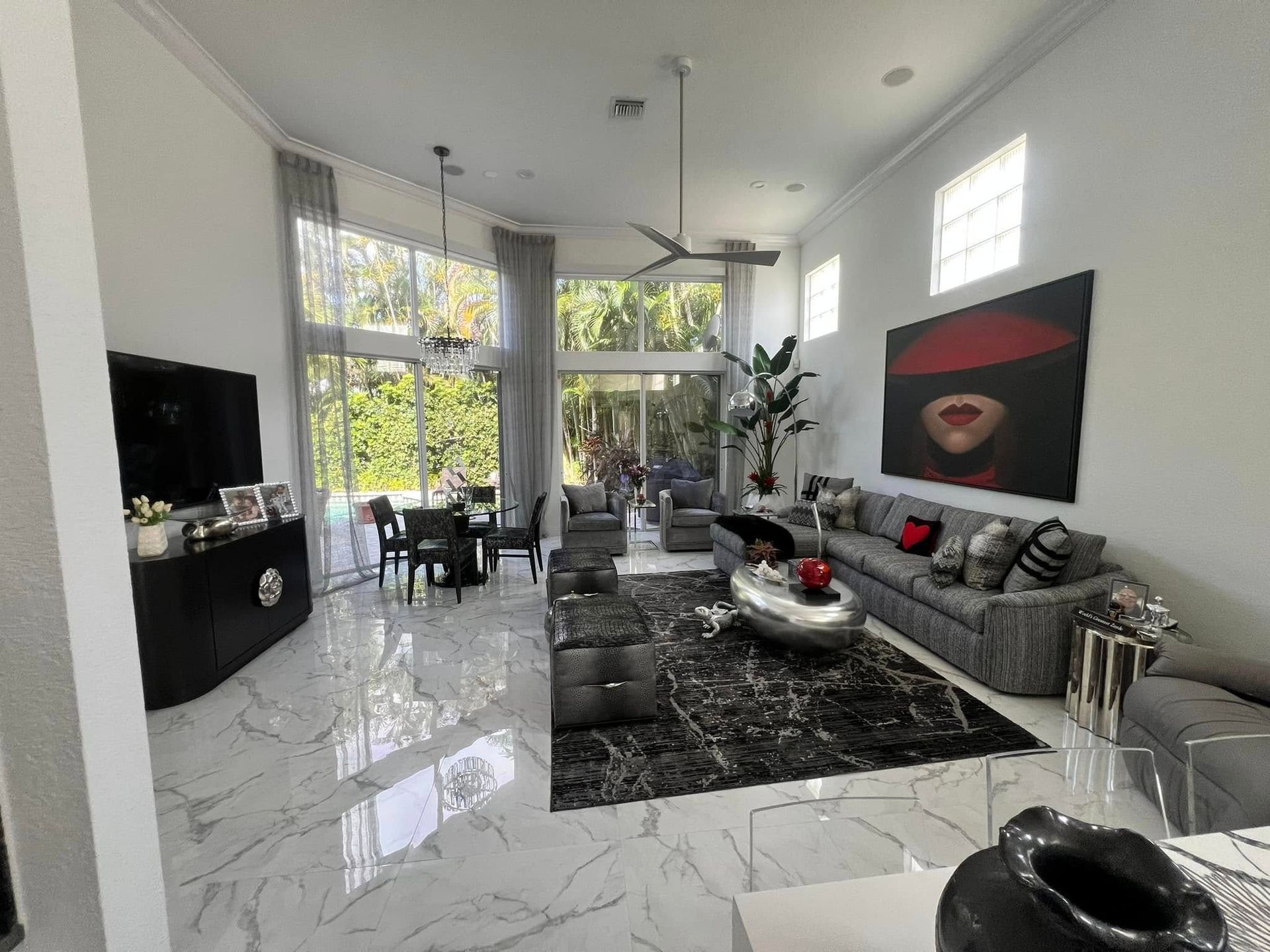 A bright, modern living room with marble floors, a large grey sofa, a black rug, and high windows overlooking trees.