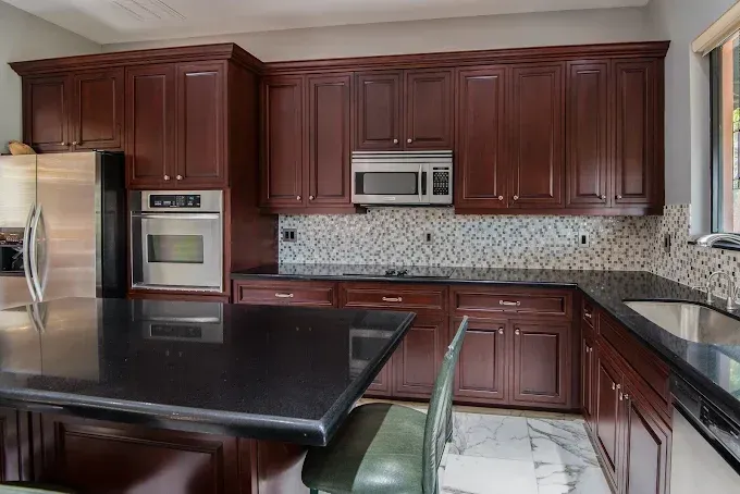 A kitchen with dark cherry wood cabinets, black granite countertops, stainless steel appliances, and mosaic tile backsplash.