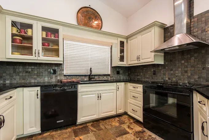 A bright kitchen with light cabinets, a black dishwasher and stove, dark tile backsplash, and a round wall decoration.