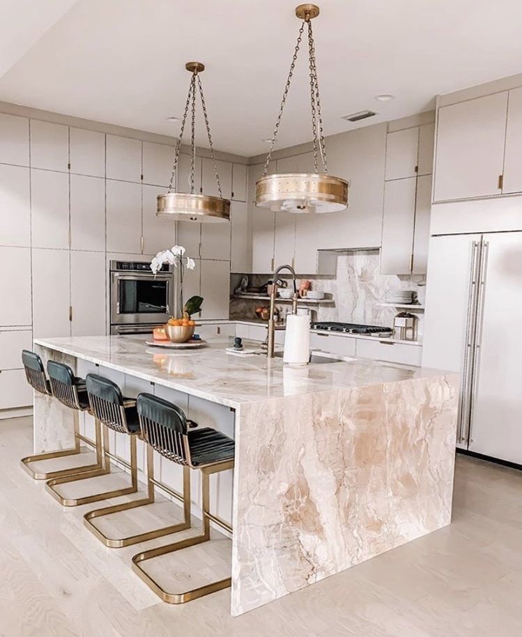 Modern kitchen with a large marble waterfall island, gold-accented bar stools, matching pendant lights, and light cabinetry.