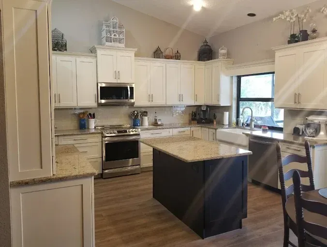 A modern kitchen with off-white cabinetry, a dark central island, granite countertops, and light wood-look flooring.