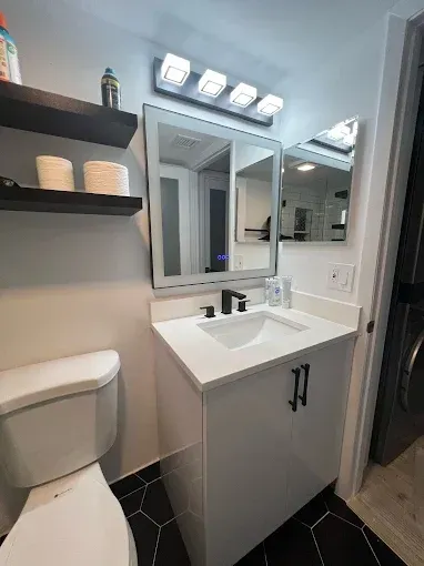 A modern bathroom with a white vanity, black hardware, square light fixture, and black hexagonal floor tiles.