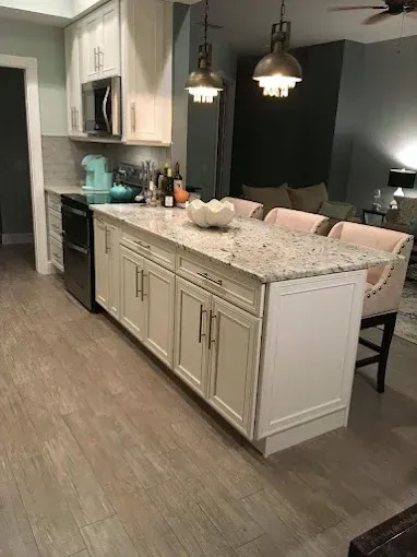 A kitchen with a large granite-topped island, white cabinetry, wood-look tile floors, and pendant lighting.