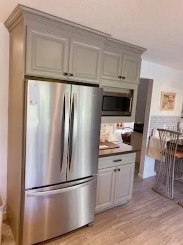 A stainless steel refrigerator sits next to grey cabinets, with a built-in microwave and base cabinets in a kitchen.