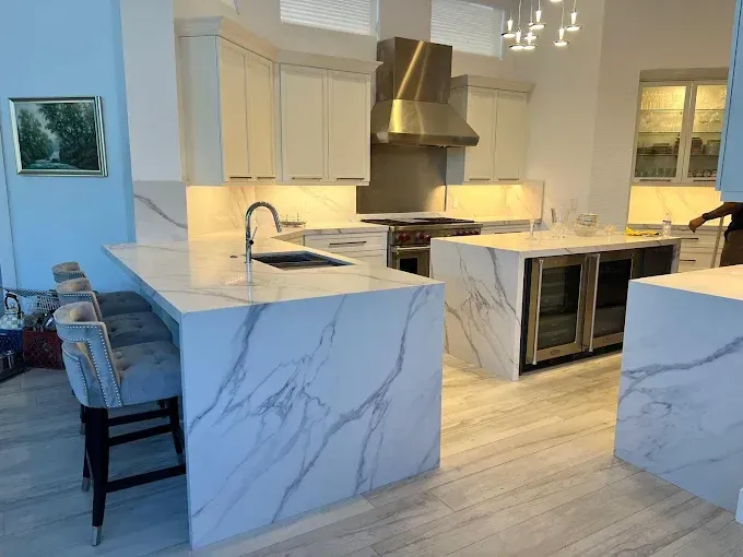 A modern kitchen with marble-topped islands, light cabinetry, stainless steel appliances, and gray upholstered stools.