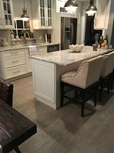 A well-lit kitchen features white cabinets, granite countertops, an island with two beige bar stools, and wood-look flooring.