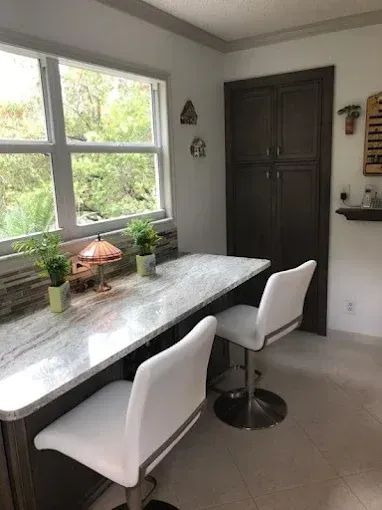 A counter with a marble-patterned top and two white bar stools sits near a window in a kitchen with dark wood cabinets.