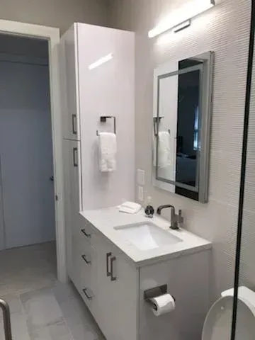 Modern white bathroom vanity with a tall storage cabinet, sink, mirror, and toilet in a bright, minimalist setting.