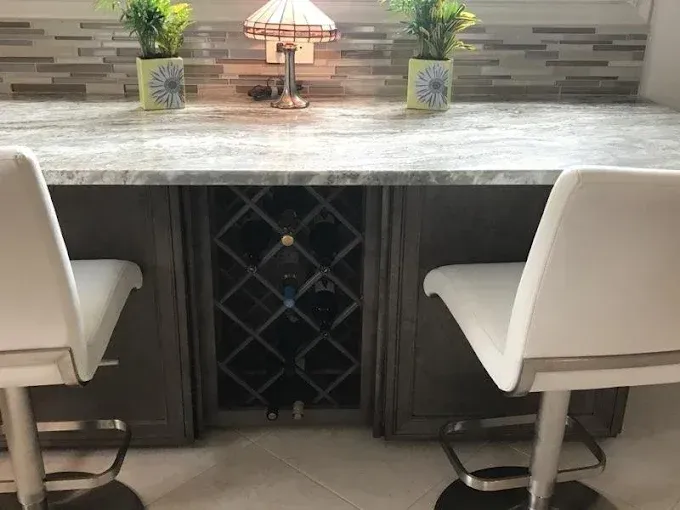 A home bar with white stools, a marble countertop, a tiled backsplash, and a wine rack containing several bottles.