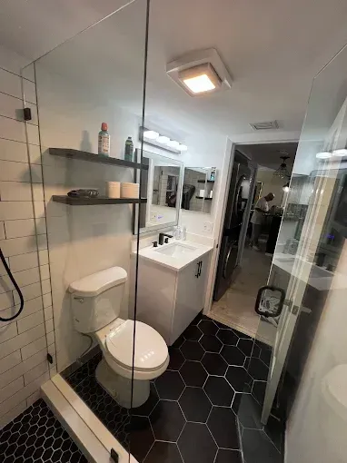 A bathroom with white subway tile, black hexagonal floor tiles, a glass shower panel, white vanity, and floating shelves.