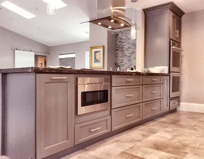 A modern kitchen island featuring grey cabinets, a built-in microwave, and drawers, with a double oven nearby.