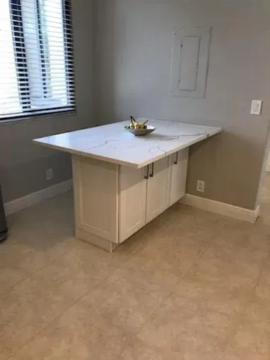 A white kitchen island with a marble-patterned countertop and cabinet storage stands in a room with light-colored flooring.